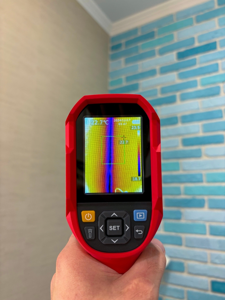 A plumber wearing blue gloves holds a digital thermal imaging camera.
